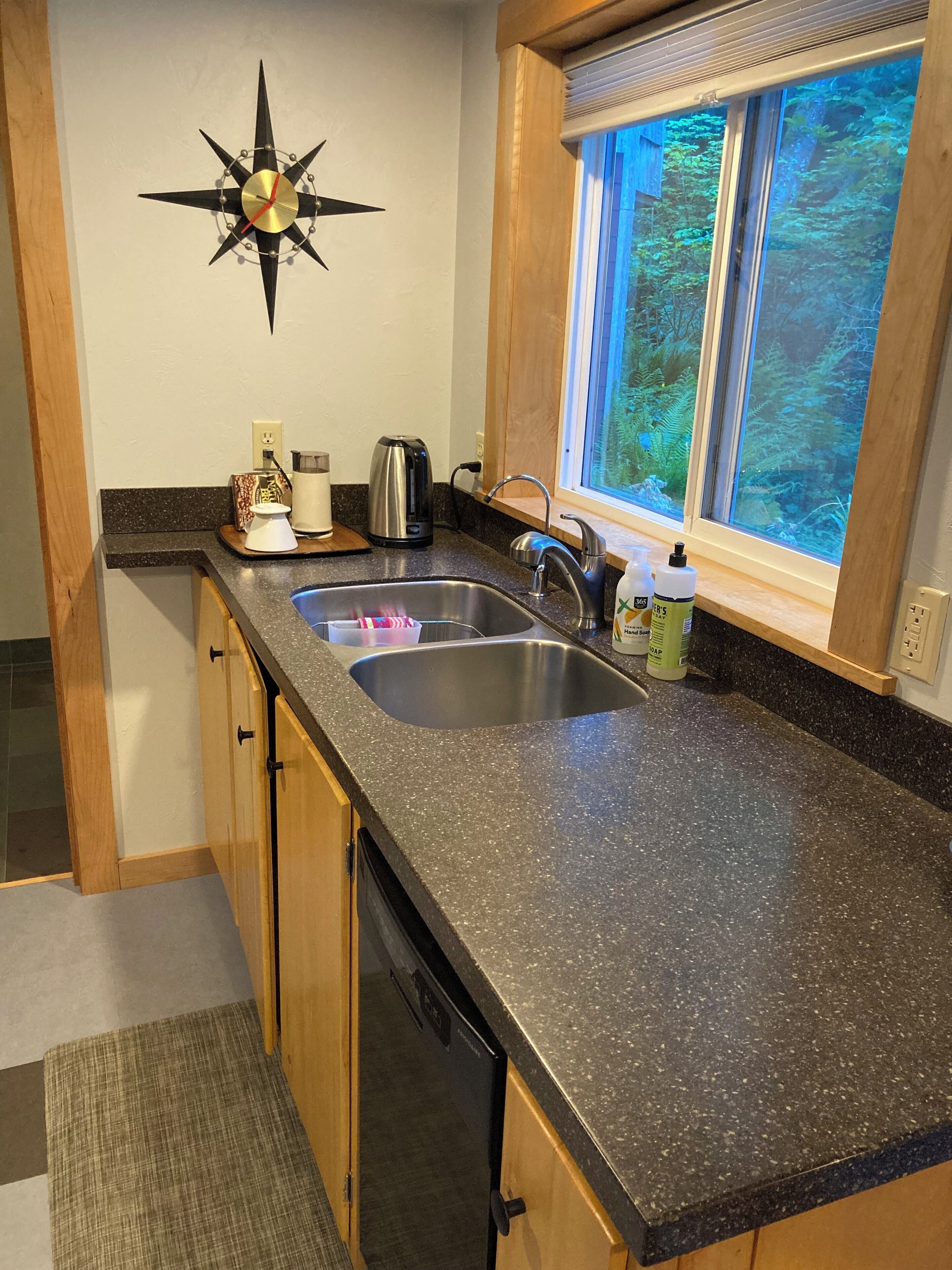 A kitchen with a granite countertop, stainless steel sink, wooden cabinets, a black clock, and a window with a view of trees.