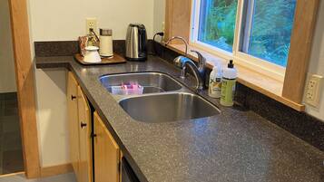 A kitchen with a granite countertop, stainless steel sink, wooden cabinets, a black clock, and a window with a view of trees.