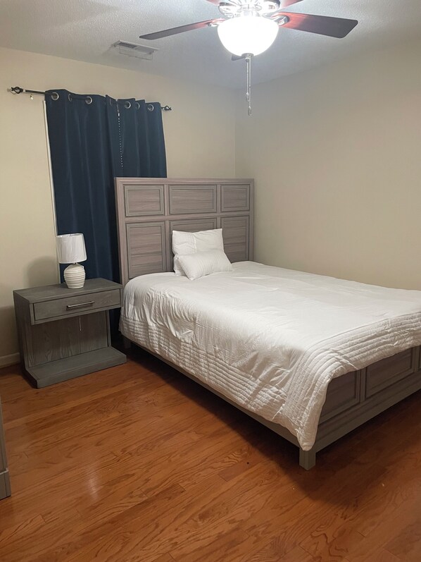 A bedroom with a bed, a nightstand, a lamp, a ceiling fan, and a window with curtains.