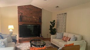 Un salon avec une cheminée en briques, un grand téléviseur à écran plat, un canapé blanc et un fauteuil marron.