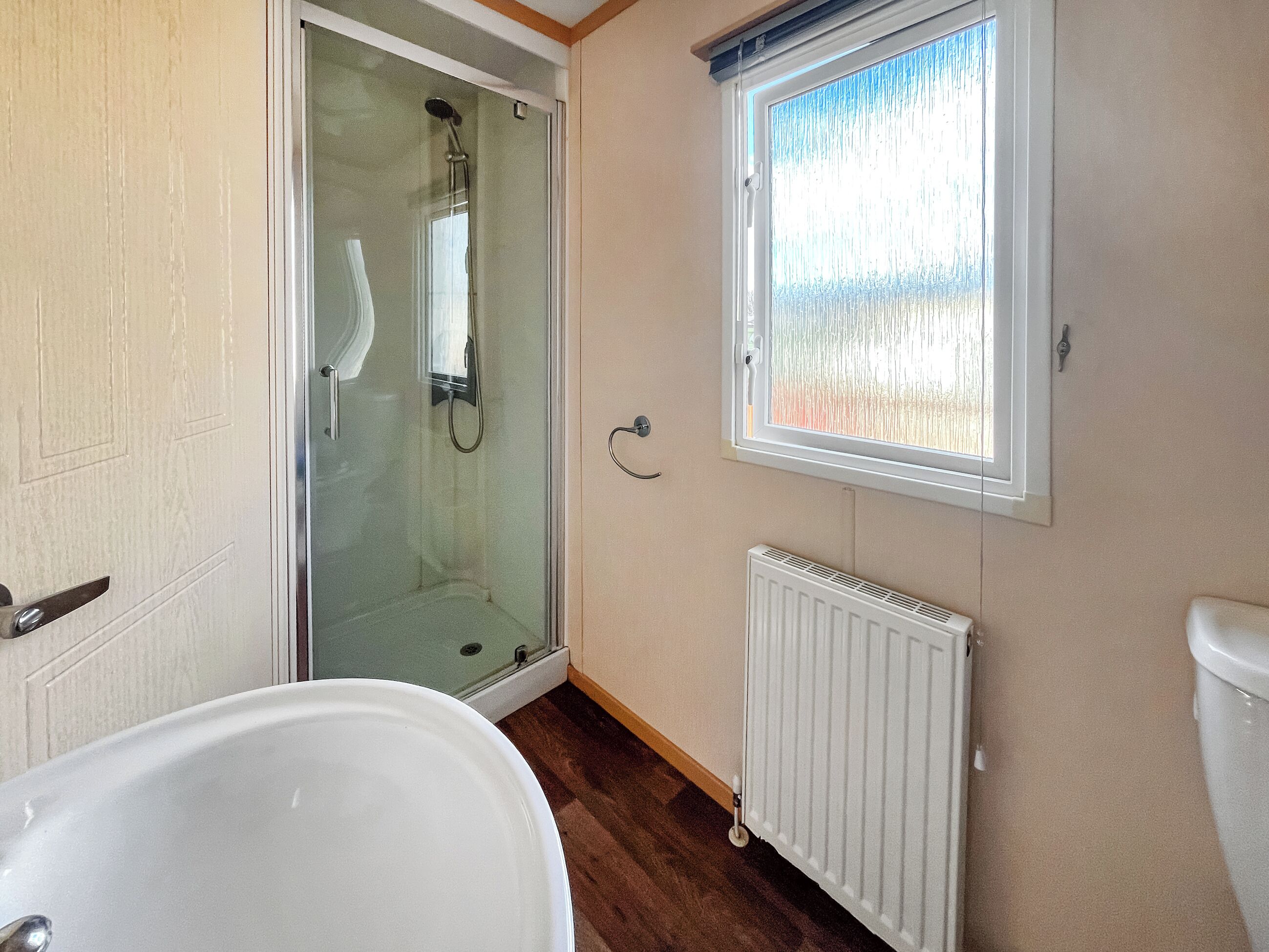 A bathroom with a glass shower enclosure, a white bathtub, a window with frosted glass, and a white radiator.