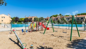 A playground with swings and slides, a pool, and a building in the background. - Family Vacation Apartment With Swimming Pools (San Michele al Tagliamento)