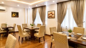Une salle à manger d’hôtel avec une table centrale, entourée de chaises, et deux fenêtres avec des rideaux.
