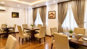 A hotel dining room with a central table, surrounded by chairs, and two windows with curtains. - Hotel JMD - Managed by Kian (New Delhi)
