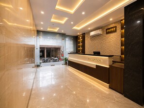 A modern reception area with a marble floor, a reception desk labeled 'RECEPTION,' and a wall-mounted air conditioning unit. - Hotel JMD - Managed by Kian (New Delhi)