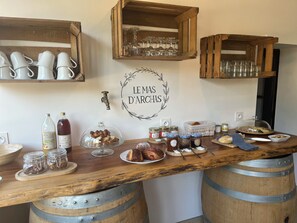 Un coin bar rustique avec des étagères en bois où sont posées des tasses et des verres blancs, un comptoir en bois sur lequel on trouve des bocaux de noix, une bouteille de vin et une enseigne indiquant « LE MAS D'ARCHES ».