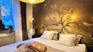 Un lit bien fait, avec du linge de lit blanc et plusieurs oreillers, un mur décoratif orné d’un motif floral et une fenêtre donnant sur des arbres.