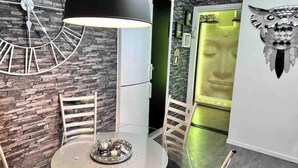 Un espace repas moderne avec une table ronde, deux chaises, une horloge murale décorative et un grand luminaire suspendu noir.