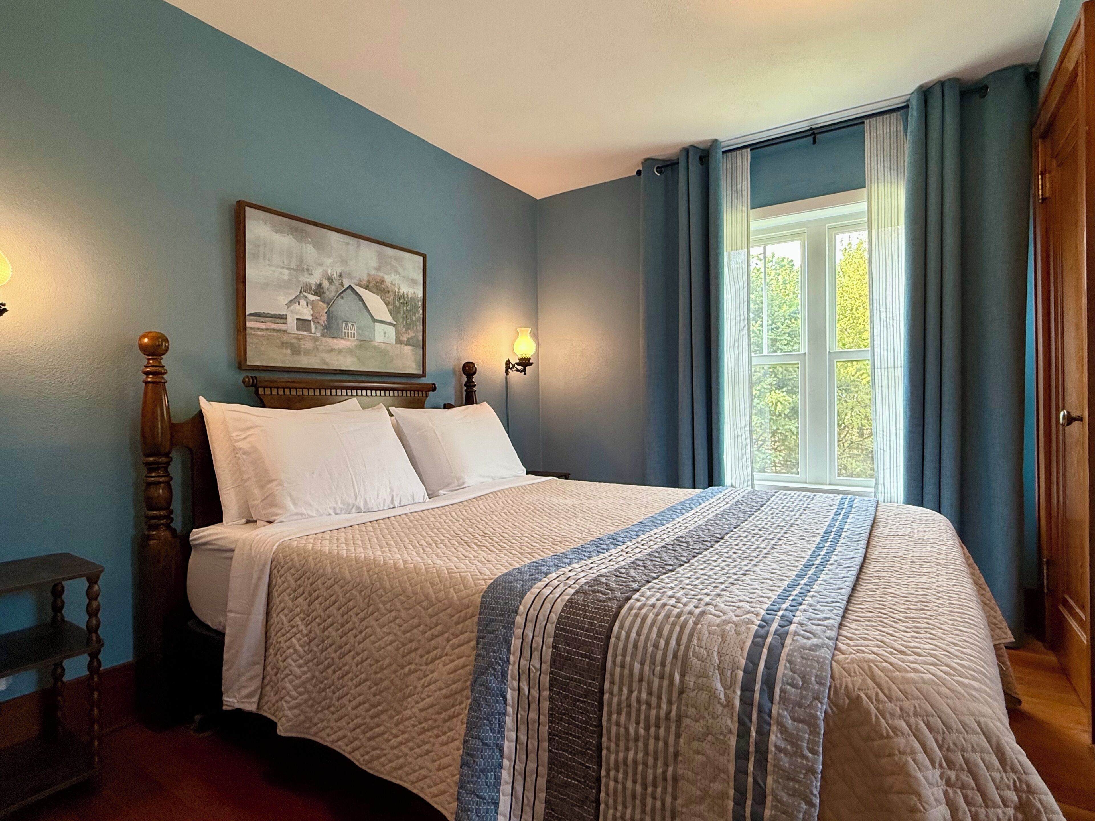 A bedroom with a large bed, a painting on the wall, and a window with curtains.