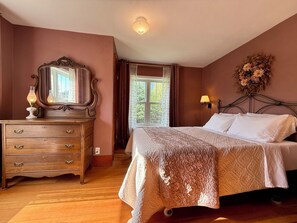 A bedroom with a wooden dresser, a large mirror, a bed with white linens, a wall-mounted lamp, and a window with curtains.