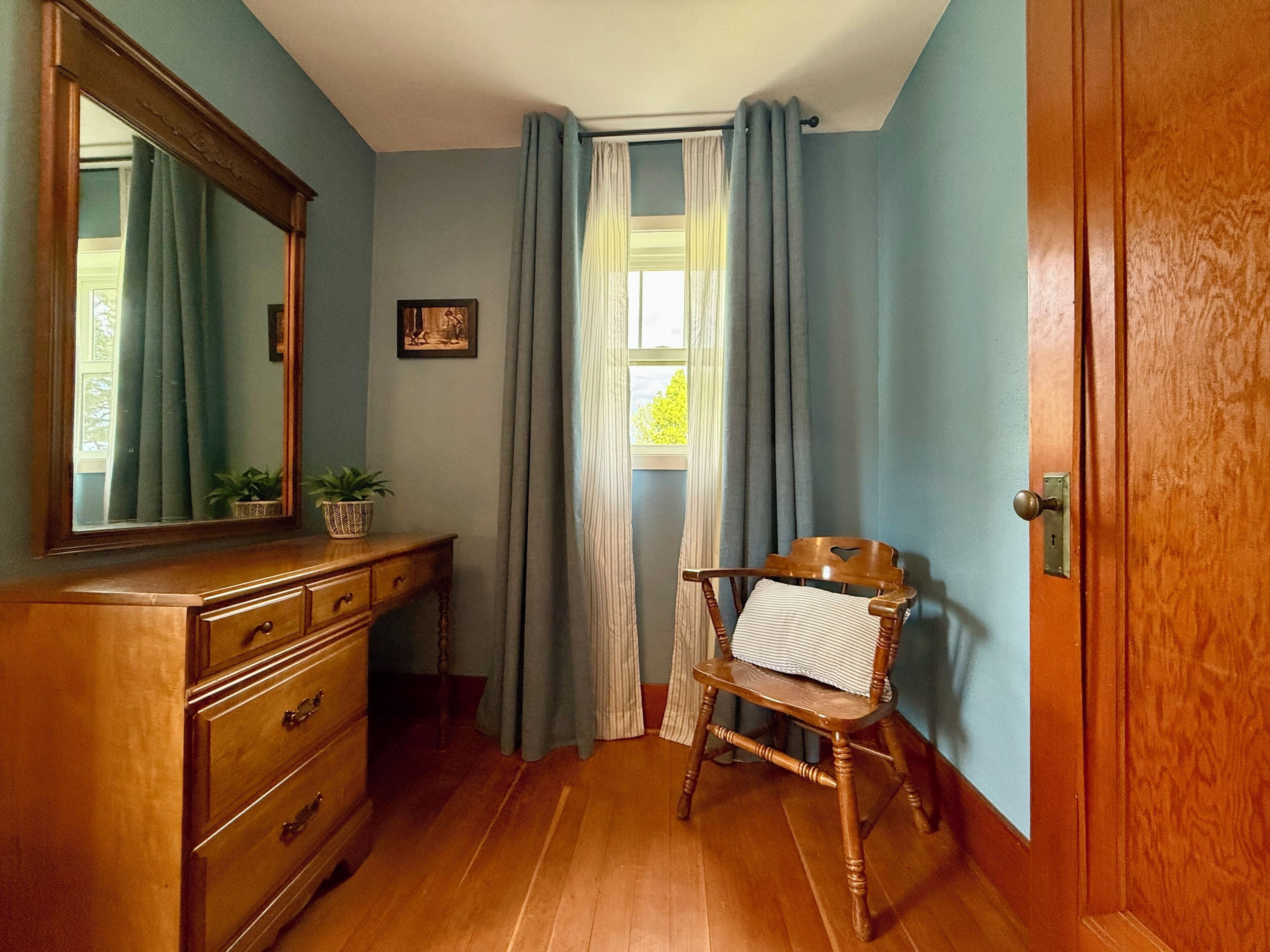 A room with a wooden dresser, a chair, a window with curtains, and a door.