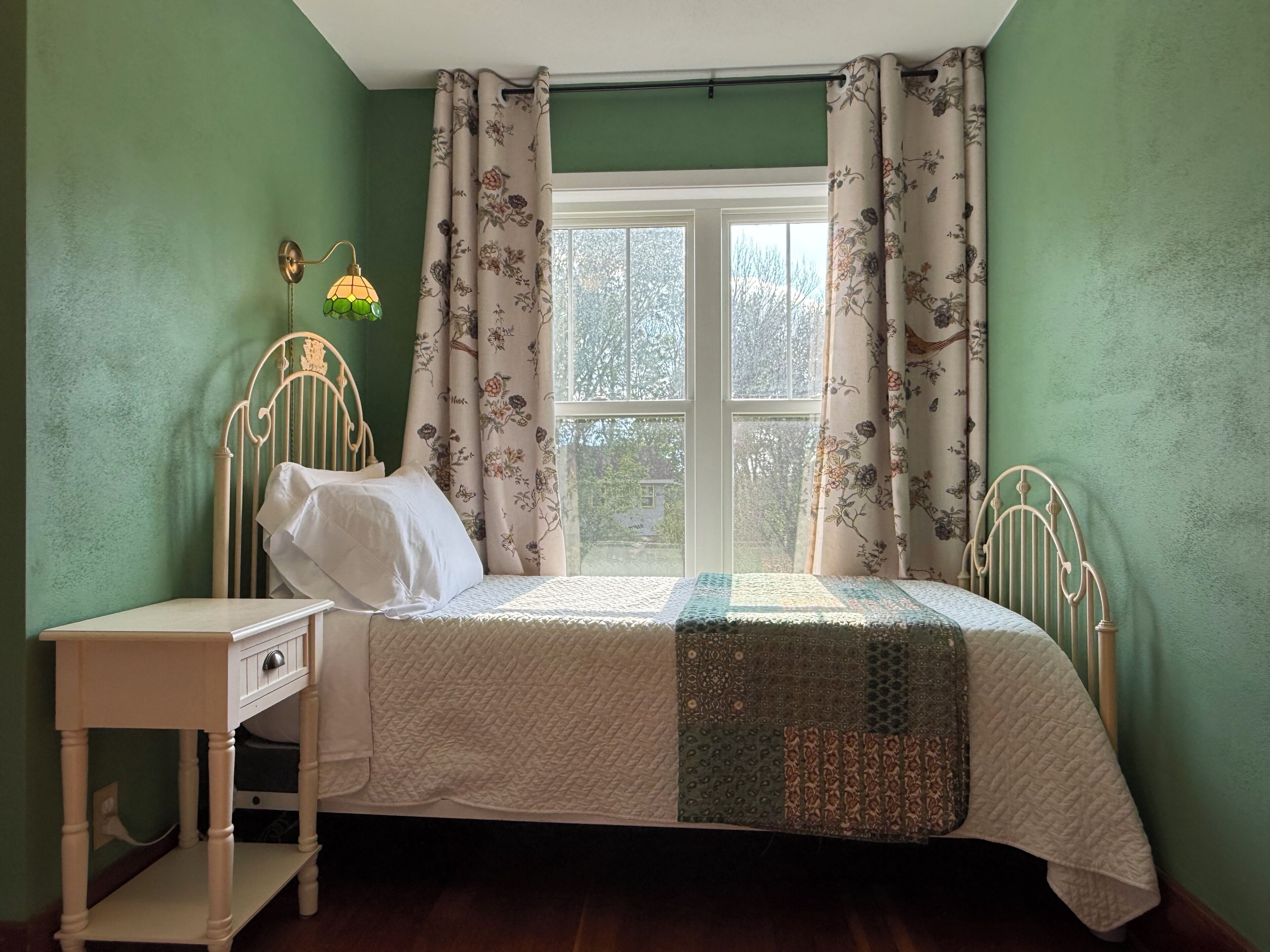 A single bed with a quilt, a bedside table, a wall-mounted lamp, and a window with floral curtains.