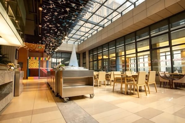 A spacious lobby with a high ceiling, a central serving cart, and a large glass wall providing a view of the outside.