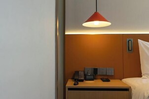 Habitación de hotel con una cama, una mesita de noche con teléfono y control remoto, una lámpara colgante roja y un panel en la pared con interruptor de luz.