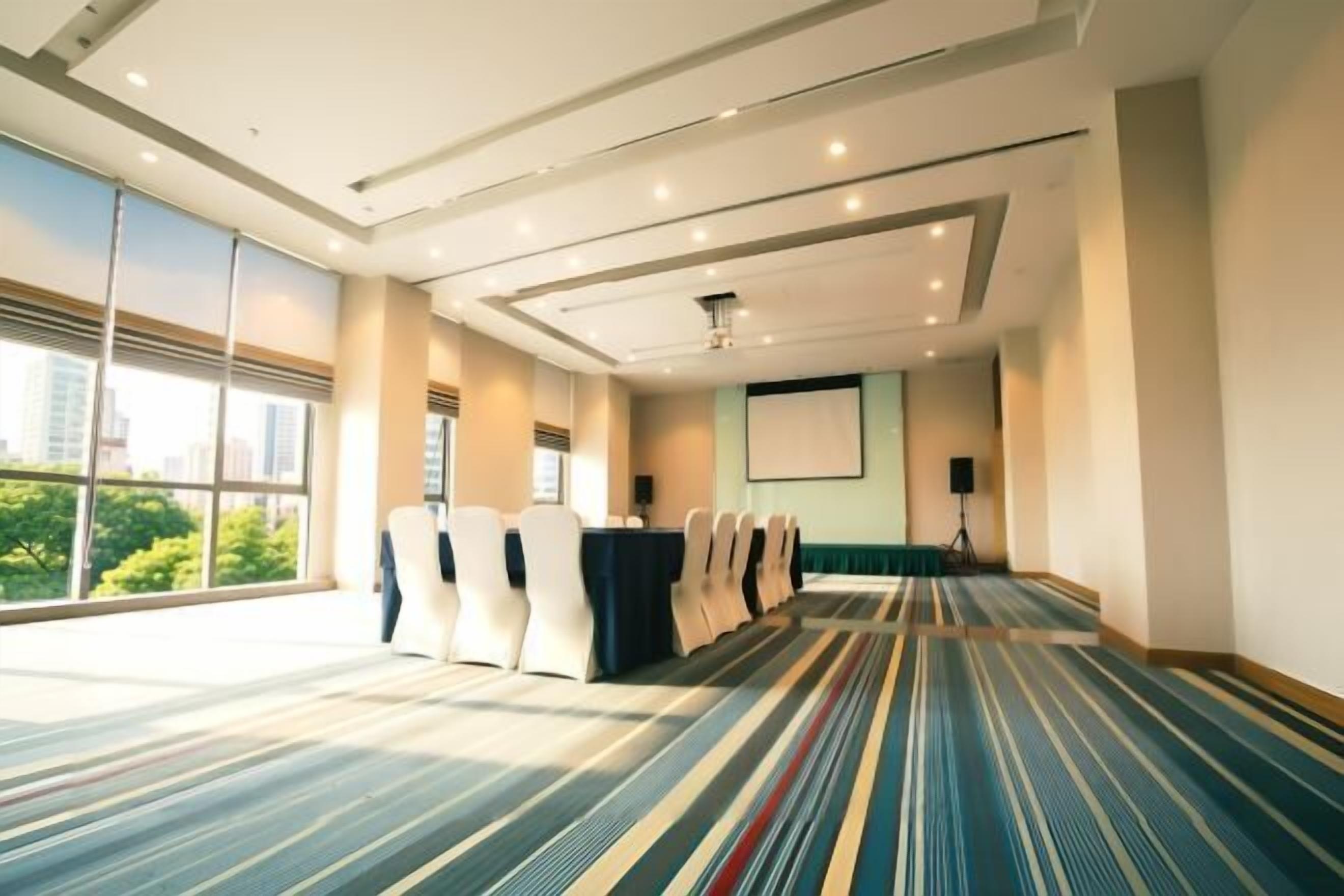 Ein Konferenzraum mit einem langen Tisch, Stühlen, einer Projektionsleinwand und großen Fenstern.