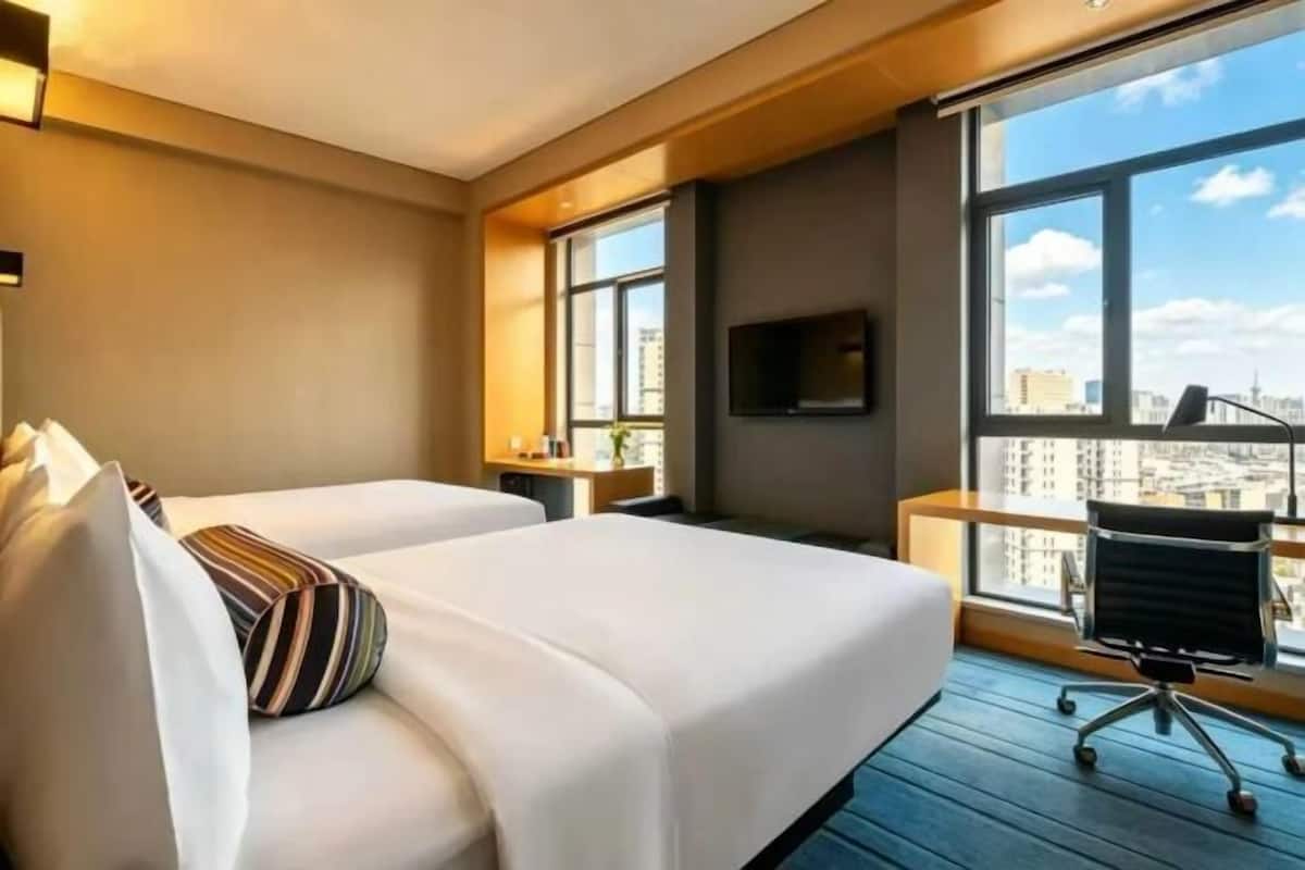 Une chambre dâhĂŽtel avec deux lits, un bureau, une chaise et une grande fenĂȘtre donnant sur la ville.