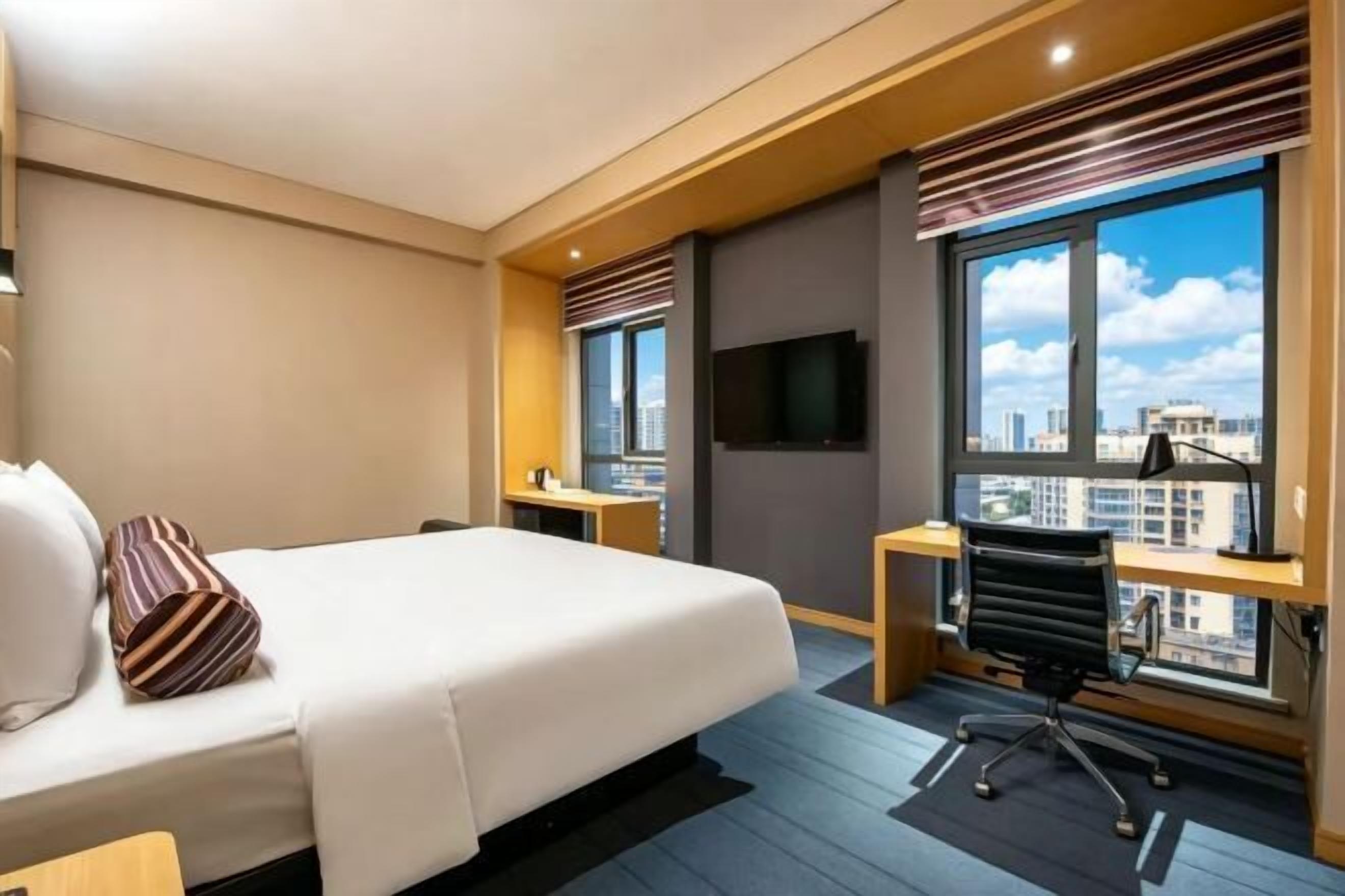 Habitación de hotel con una cama grande, un escritorio, una silla, un televisor y una ventana con vistas a la ciudad.