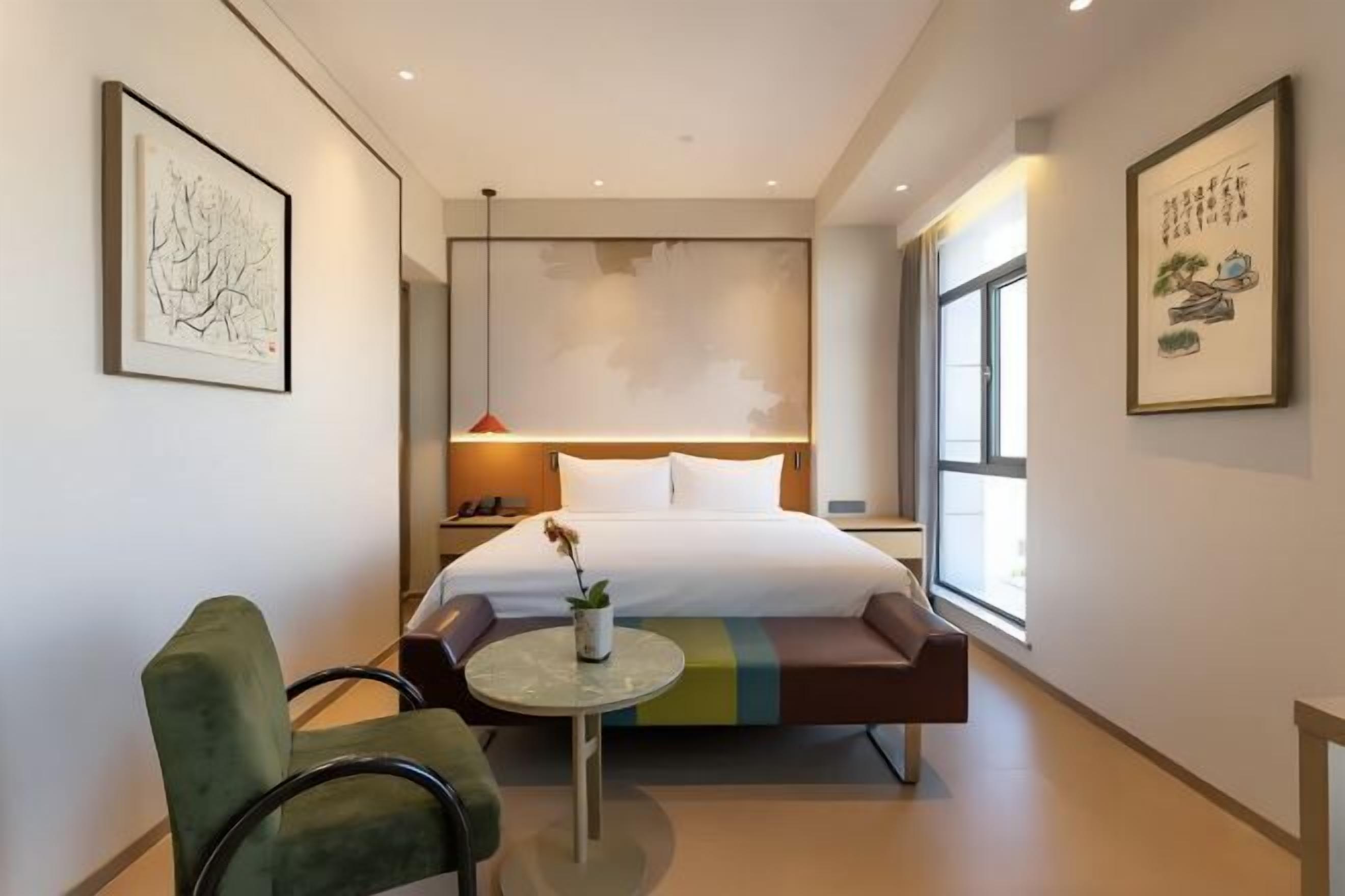 A hotel room with a bed, a small table with a plant, a green armchair, and artwork on the walls.