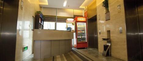 A modern hotel lobby with a reception desk, vending machine, and elevators.