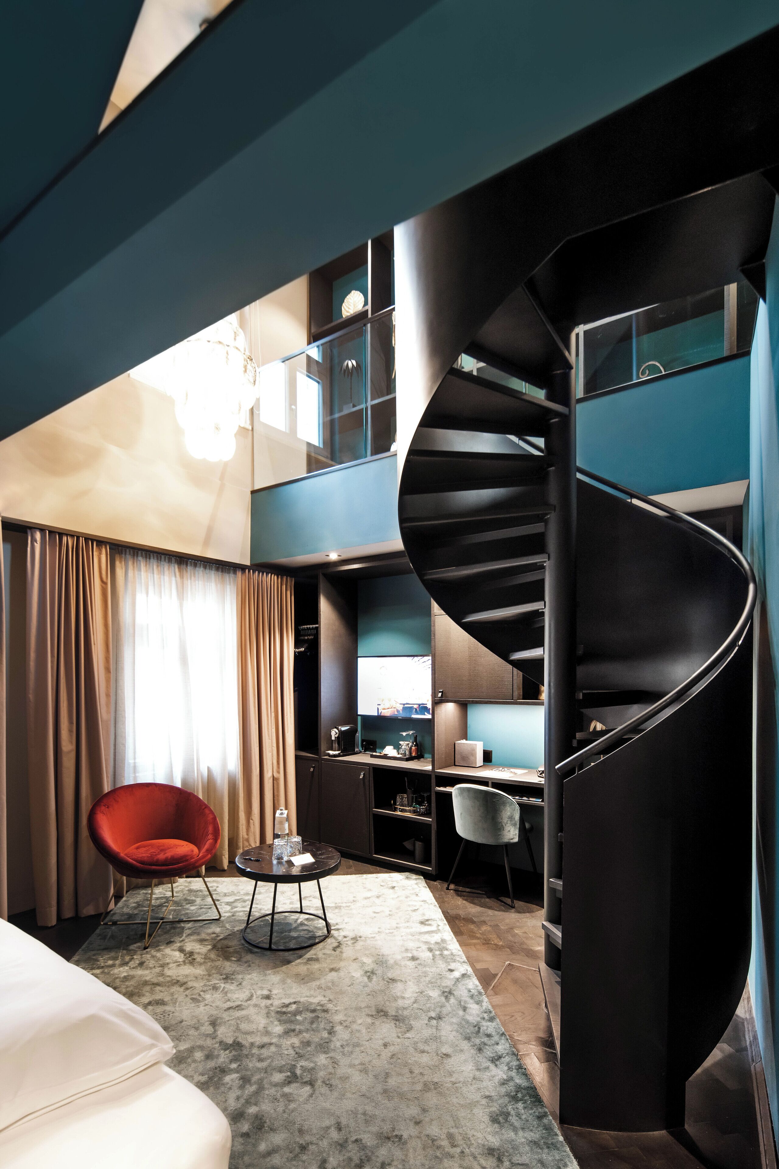 A modern hotel room with a spiral staircase, a red chair, a small table, and a large bed.
