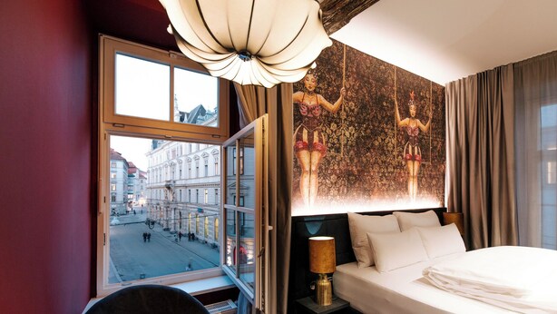 A hotel room with a large bed, a chair, a nightstand with a lamp, a view of a city street through a window, and a chandelier. - Aiola Living Boutique Hotel Graz (Graz)