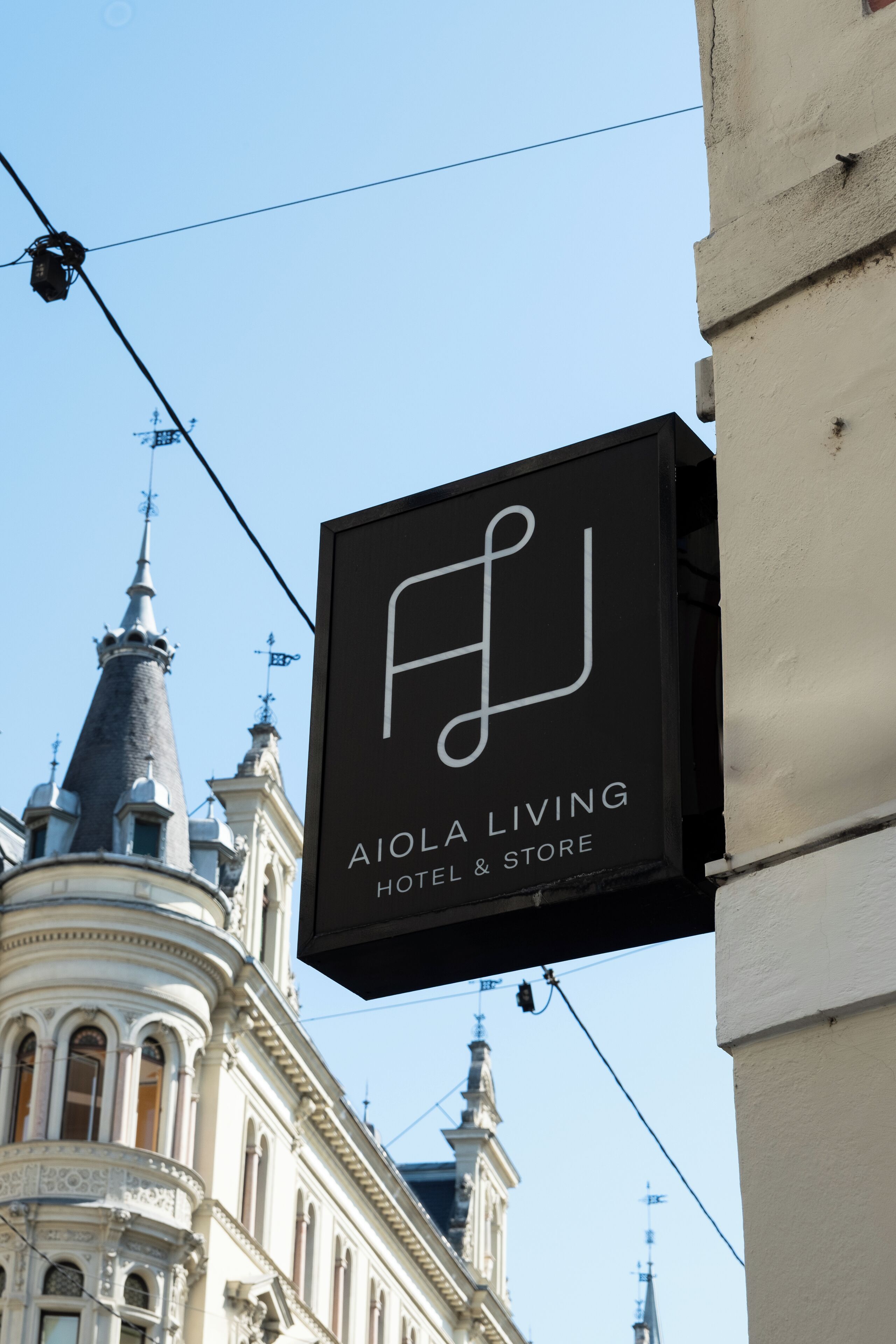 Un letrero negro con letras blancas que dice 'AIOLA LIVING HOTEL & STORE' frente a un edificio de arquitectura ornamentada.