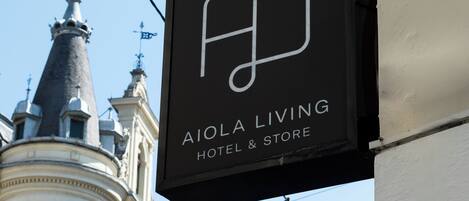 Un letrero negro con letras blancas que dice 'AIOLA LIVING HOTEL & STORE' frente a un edificio de arquitectura ornamentada.