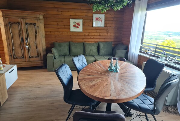 A cozy room with a wooden floor, a round wooden table with chairs, a green sofa, and a large window with a view.