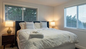 A neatly made bed with a quilted cover, two bedside lamps, a dark headboard, and a window with blinds. - Sauna, Beach, Sunset, Game Room, Peace and Quiet, Fun! (Oak Harbor)