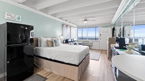 A modern room with a bed, a refrigerator, a kitchen area, and a large window offering a view. - Beachfront Studio, Sleeps 2, Pool (Panama City Beach)