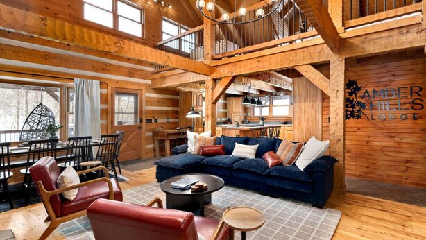 A spacious living area with a large sofa, red leather chairs, and a dining table. A kitchen is visible in the background, and the room has wooden walls and a high ceiling. - Amber Hills Lodge: Sauna,Golf Sim,Hot Tub,Theatre (Lancaster)
