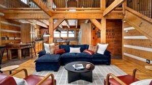 A log cabin interior with a large blue sectional sofa, leather chairs, a round coffee table, and a kitchen area visible through an open door. - Amber Hills Lodge: Sauna,Golf Sim,Hot Tub,Theatre (Lancaster)