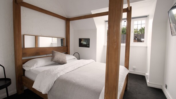 A bedroom with a wooden bed frame, a white bedspread, and a single pillow. There is a window with a view of buildings outside. - Cosy Family One Bedroom flat with private Kitchen (Sutton)