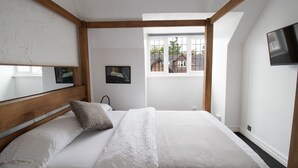 A bedroom with a wooden headboard, a bed with white linens, a window with a view of buildings, and a flat-screen TV mounted on the wall. - Cosy Family One Bedroom flat with private Kitchen (Sutton)