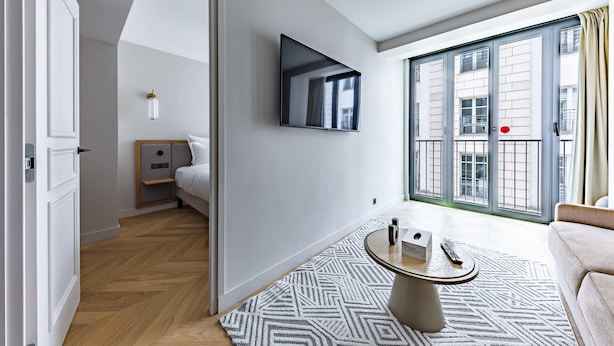 Una habitación de hotel moderna con cama, televisor montado en la pared, una mesita de noche pequeña y una mesa de centro redonda, y un ventanal con vistas a edificios.