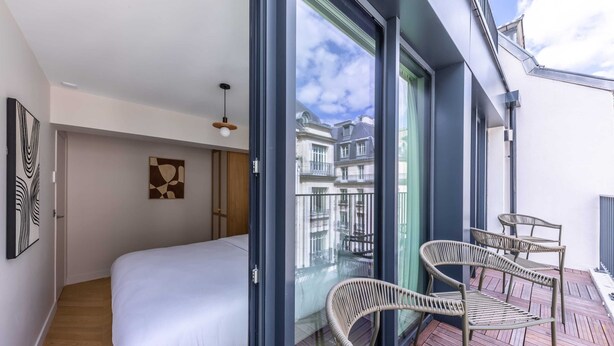A modern bedroom with a large bed, a glass door leading to a balcony with wicker chairs, and a view of neighboring buildings. - Opera - One Bedroom Balcony (Paris)