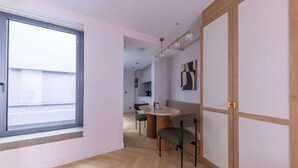 A modern room with a large window, a round table with chairs, a wooden wardrobe, and a kitchen in the background.