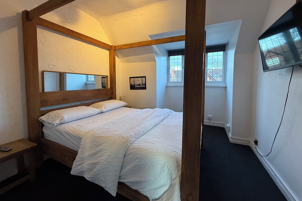 A four-poster bed with a wooden headboard, a bedside table, a flat-screen TV mounted on the wall, and a window with a view of the outside.