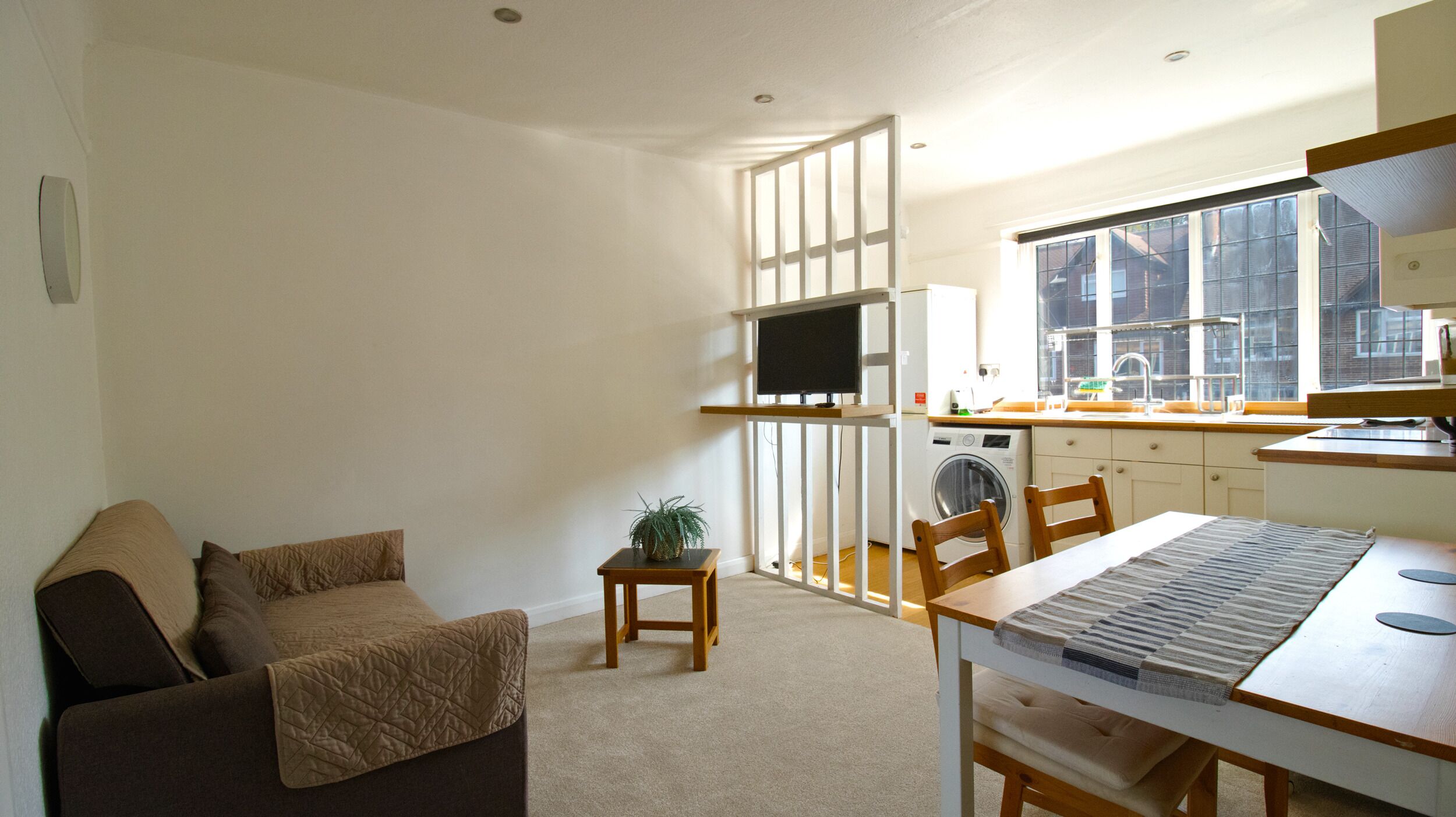 Un espacio habitable compacto con sofá, mesa de comedor, mesita auxiliar con planta, lavadora y ventana con vistas a edificios.