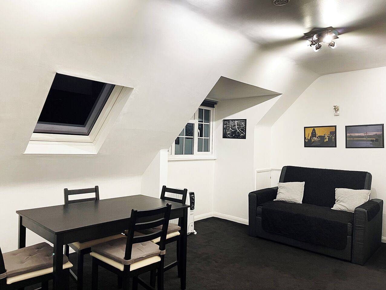 Quarto com mesa de jantar, cadeiras, sofá preto, janela e quadros na parede.
