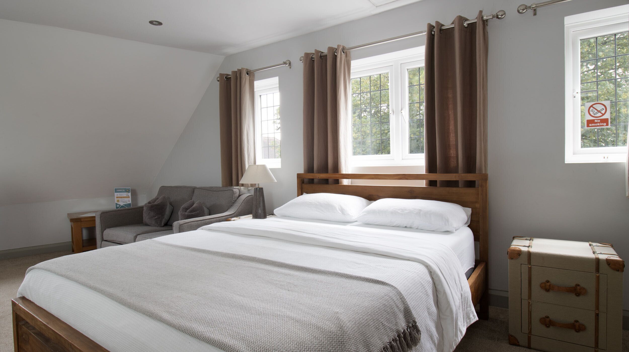 A bedroom with a bed, a nightstand, a chair, a suitcase, and a window with curtains.