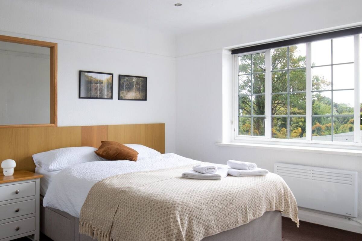 A bedroom with a bed, a nightstand, a window with a view, and two framed pictures on the wall.
