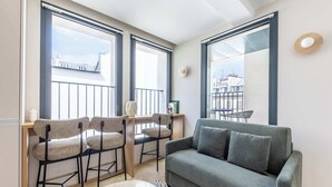 A modern living room with a grey sofa, two bar stools, a round table, and large windows offering a view of buildings.