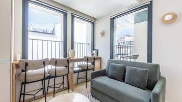 Una sala moderna con un sofá gris, dos taburetes altos, una mesa redonda y ventanas amplias que ofrecen vistas a edificios.
