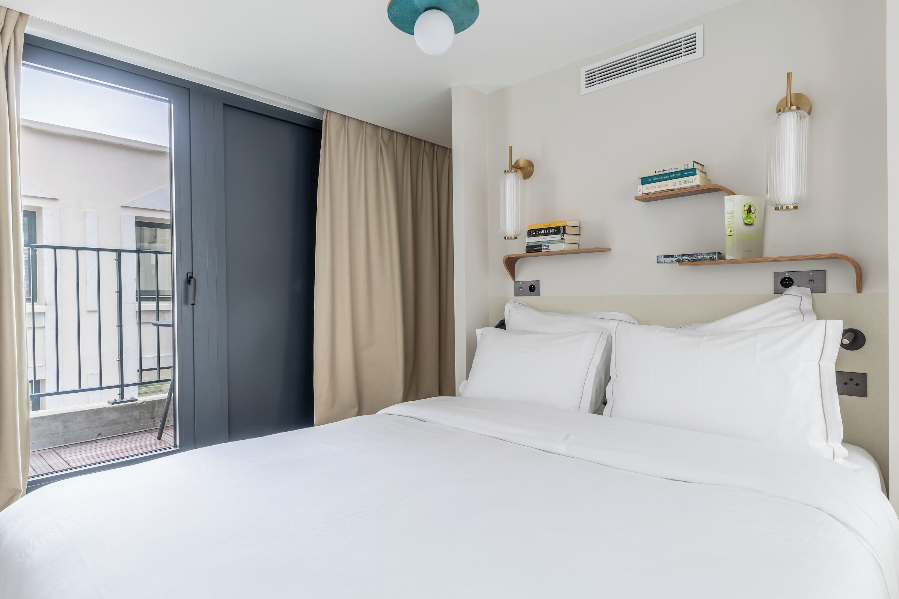 A hotel room with a bed, a sliding door to a balcony, a wall-mounted shelf with books and a lamp, and a ceiling vent.