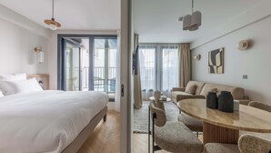 A hotel room with a bed, a dining table with chairs, a sofa, and a large window with curtains.