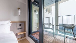 A modern bedroom with a bed, a bedside table, a window with a view of a balcony with chairs and a railing.