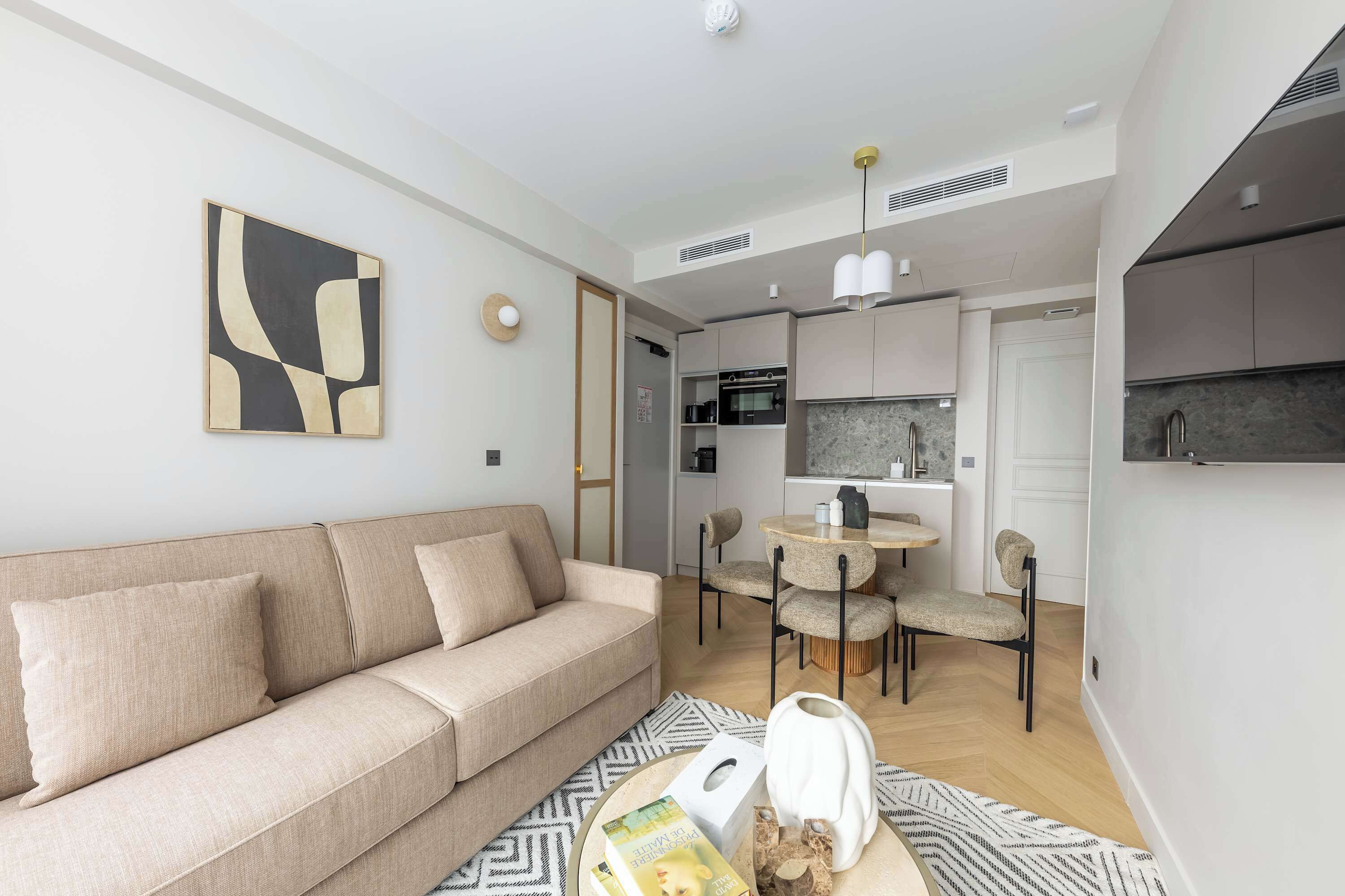 A modern living room with a sofa, a dining area with chairs, a kitchen with cabinets, and a ceiling light.