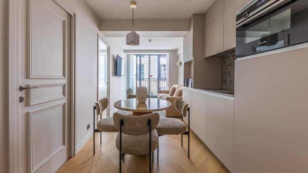 Une cuisine moderne dotée d’un four encastré, un coin repas avec une table ronde et des chaises, et une grande fenêtre laissant entrer la lumière naturelle.