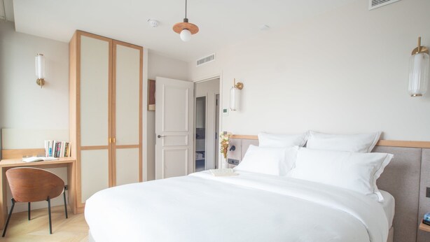 A hotel room with a large bed, a desk with a chair, a wardrobe, and a bedside table with a lamp.
